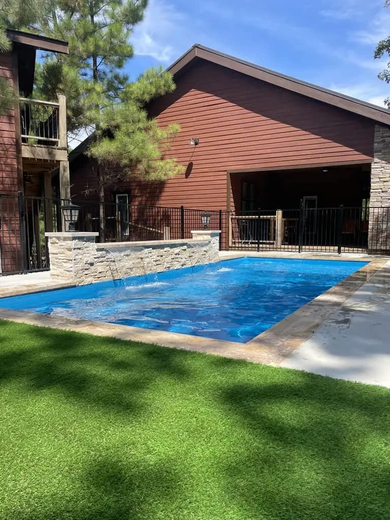 A sparkling blue swimming pool surrounded by lush artificial grass and a wooden cabin, offering a tranquil outdoor relaxation space.